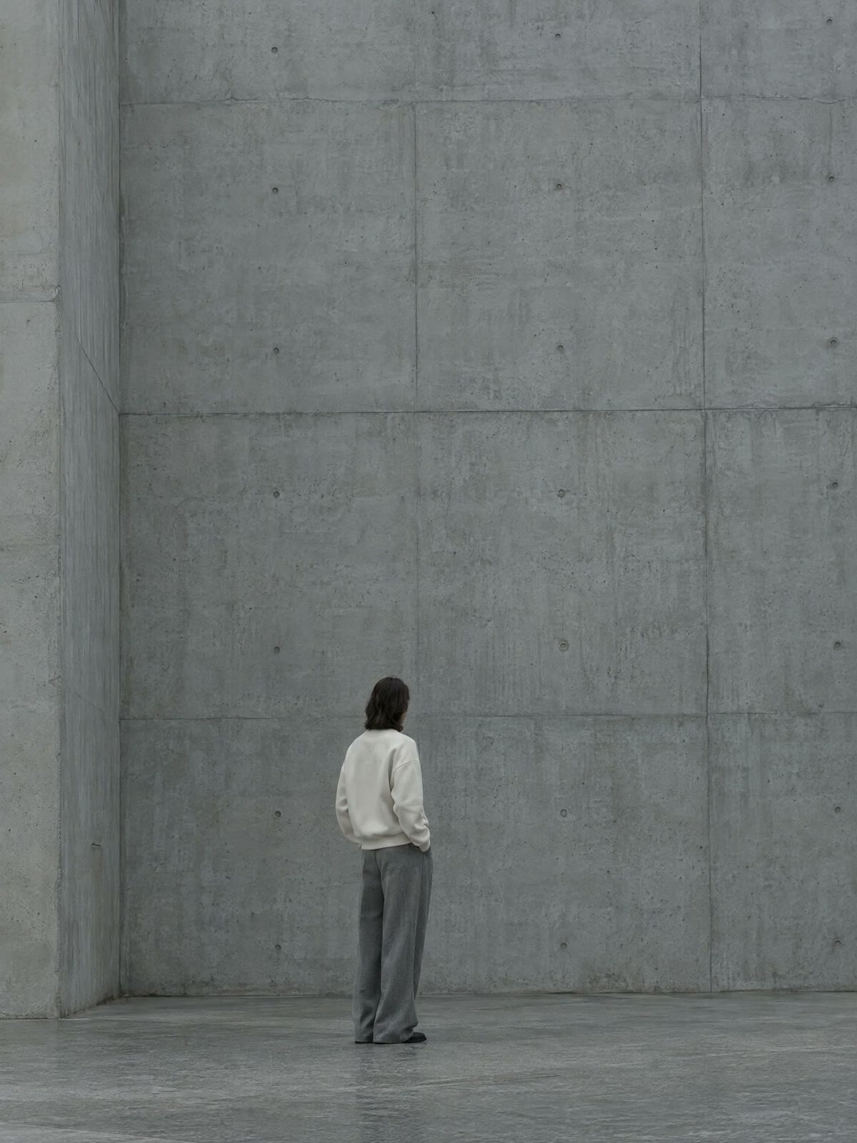 A figure in a bone crewneck standing against a poured concrete wall.